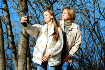 Fototapeta premium Mature mother and adult daughter are doing selfie by mobile phone in summer park, near lake.