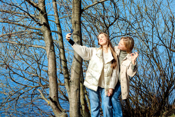 Mature mother and adult daughter are doing selfie by mobile phone in summer park, near lake.