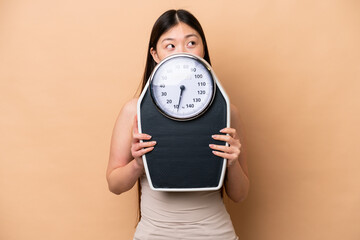 Young Chinese woman isolated on beige background with weighing machine and hiding behind it