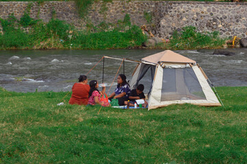 Two mothers sit conversing in front of the tent as the daughter drinks and son eats during their...