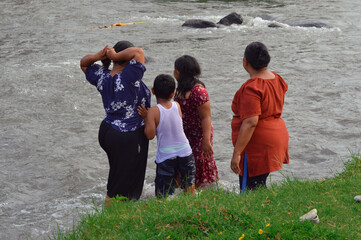 The group of two women, a boy, and a girl stand side-by-side on the riverbank, gazing into the...