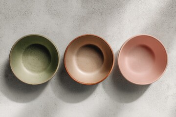Three empty, shallow bowls in muted green, beige, and pink, arranged in a row on a light gray surface with subtle shadows