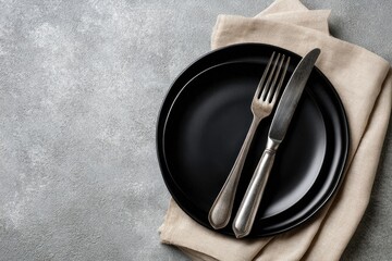 Two black plates, silver fork and knife rest on a beige linen napkin atop a grey textured surface