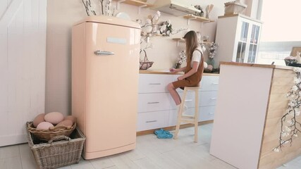 Young girl writing at a kitchen counter in a bright room with a retro fridge and Easter decorations. Cozy domestic scene.
