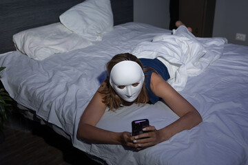 In a dimly lit bedroom, a person wearing a white mask lies on their stomach on a bed while intently...