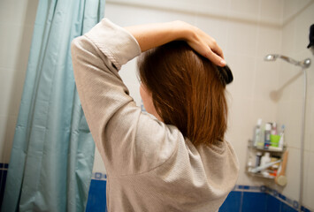 Obraz premium Person seen from behind brushing brown hair in bathroom. Daily self-care and hygiene routine. Features beige sweater, blue curtain, toiletries. Focus on hair care inside.