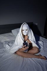 A person sits on a bed, covered in white fabric, holding a smartphone. They wear a white mask,...