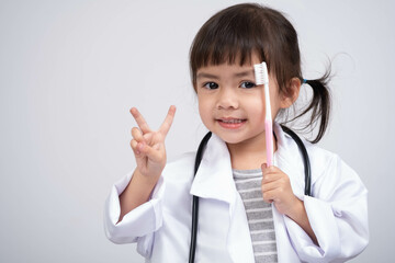 A cheerful young girl in a doctor's coat holds a toothbrush while flashing a peace sign, promoting...