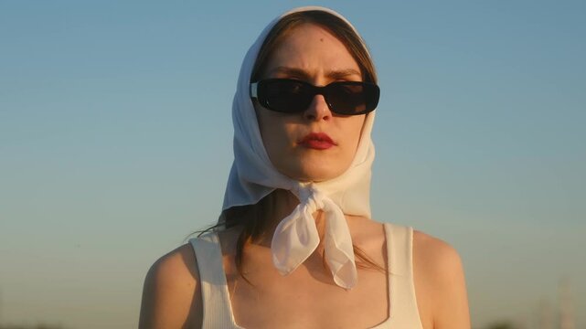 Fashion model posing confidently in white headscarf and sunglasses, showcasing stylish expressions against clear blue summer sky, embodying contemporary elegance and individual beauty