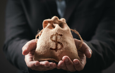 A person holds a small burlap sack with a dollar sign, symbolizing money, wealth, or financial...