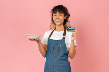 Beautiful Asian women wear apron and holding white dishes with credit card on pink background.