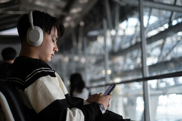 A young man wearing headphones sits in an airport, focused on his smartphone amidst a modern...