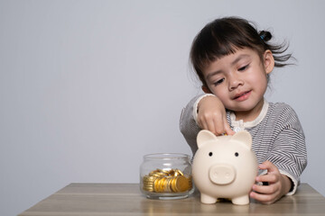 A young girl happily deposits coins into a piggy bank, promoting the concept of saving and...