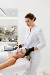 Mature woman receiving anti-aging skin treatment lying in cosmetic clinic. Cosmetologist performing photorejuvenation face procedure using professional aesthetic laser machine, skincare and wellness