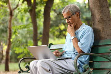 Indian senior man working laptop in park. old man wearing blue t-shirts and eye wear glasses while...