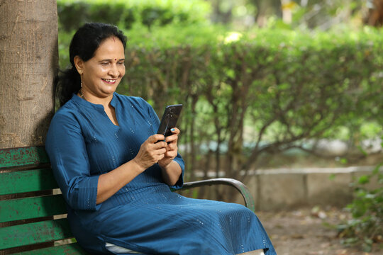Happy Indian senior woman watching video or social media on smart phone while sitting on bench at park outdoor, Mature old people using technology.