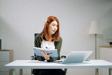 Young Asian woman in office focusing on laptop and writing in notebook. Concept of productivity,...