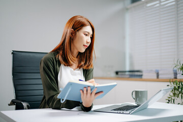 Young Asian woman in office focusing on laptop and writing in notebook. Concept of productivity,...