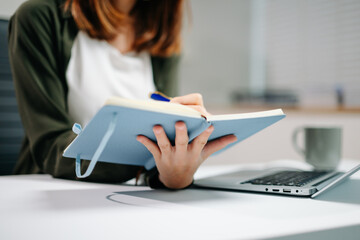 Young Asian woman in office focusing on laptop and writing in notebook. Concept of productivity,...