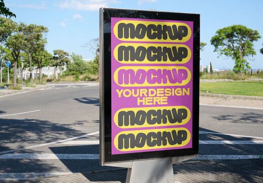 A blank urban panel mockup stands at a street intersection, surrounded by greenery and clear blue skies, ready for advertisement placement.