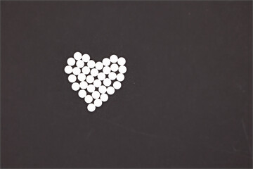 A Heart Shape Carefully Arranged From Small Round White Pills Or Tablets On A Plain Black...