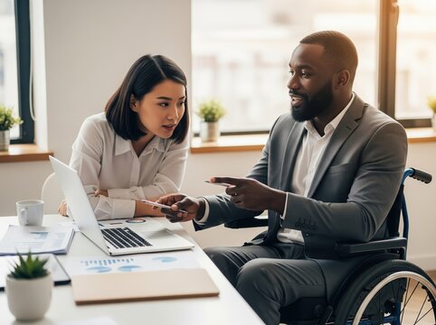 Handicapped man in wheelchair at desk shares creative project ideas with female coworker during casual business meeting in workplace environment - Powered by Adobe
