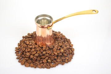 A shiny copper cezve with a brass handle alongside a pile of coffee beans on a bright white...