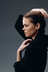 serious woman posing in black jacket with hands near head in studio lighting, showcasing beauty and...