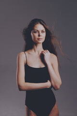 thoughtful woman in black bodysuit posing with natural makeup and loose hair against gray...