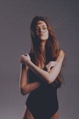 young woman with long brown hair wearing black bodysuit posing with crossed arms against a neutral...