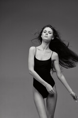 Fototapeta premium black and white woman wearing a black bodysuit poses confidently with flowing hair against a plain gray background, showcasing elegance and minimalistic style.