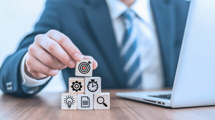 A professional business photo showing a hand in a dark navy suit and blue striped tie placing wooden blocks on a wooden desk next to a silver laptop