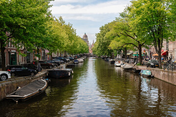 An Amsterdam canal flanked by mature trees and historic buildings, featuring various boats and parked vehicles