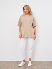 Blonde woman posing in a studio wearing a casual beige outfit with white pants
