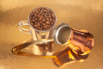 A copper coffee pot with a brass handle and a glass cup filled with coffee beans on a reflective...