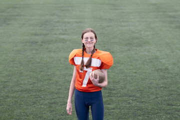 A girl wearing an orange football jersey with the number 7 stands on a grassy field. She holds a...