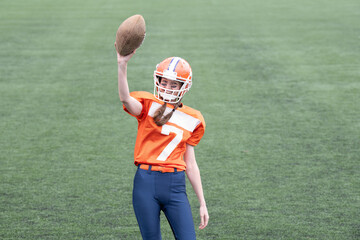 On a sunny day, a young woman wearing orange football gear holds a ball high while standing on the...
