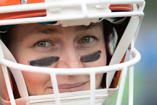 A woman is geared up in a football helmet, displaying a confident smile. She has black eye paint and is ready for the upcoming game, embodying passion for the sport - Powered by Adobe