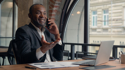 Happy smiling laughing African American positive ethnic man business employer entrepreneur smile pleasant conversation smartphone call talk mobile phone businessman guy male in office typing laptop