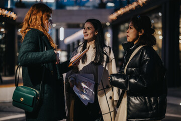 Three women engage in a conversation in a bustling urban setting, discussing documents. The lighting suggests a cold season, and their attire reflects warmth and comfort.