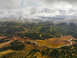Obraz premium An aerial view depicts a winding river flowing through golden-brown marshland and green forests, with snow-covered mountains in the distance