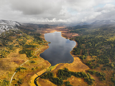 A sweeping aerial view of Yazovoe Lake, embraced by a valley of vibrant autumn foliage and distant snow-covered mountains under a cloudy sky