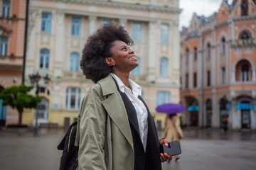 Businesswoman with afro hair, smiling and looking up while walking in the city center on a rainy...
