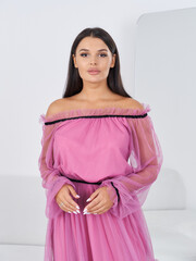 Woman with long, dark hair wears an off-shoulder pink dress with transparent sleeves. She stands...