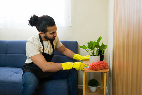 Male housekeeper cleaning furniture in living room with duster and gloves