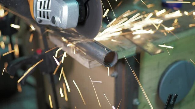 Workshop. A worker uses a angle grinder to cut a steel pipe that is fixed to a workbench with a clamp along a marked line. Close-up.