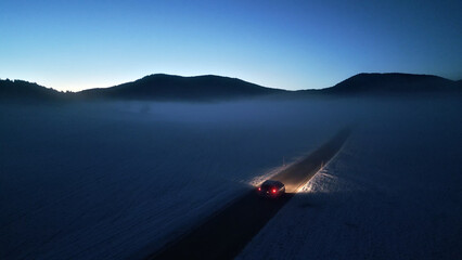 A car drives down a winding mountain road at night, its headlights illuminating the foggy, snow-covered landscape. The scene evokes a sense of adventure and the thrill of alpine driving. Aerial view. © wkproduction