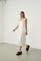 A woman in a sleeveless white dress and black platform sandals walks confidently indoors. Her hair...