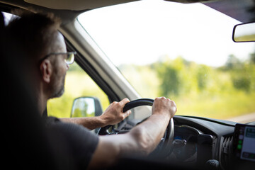 Close-up of a man driving a car on a sunny day, focusing on steering wheel and road ahead.