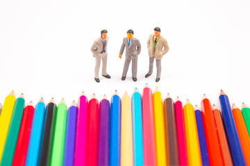 A colorful array of sharpened colored pencils contrasts with three tiny businessman figurines on a...